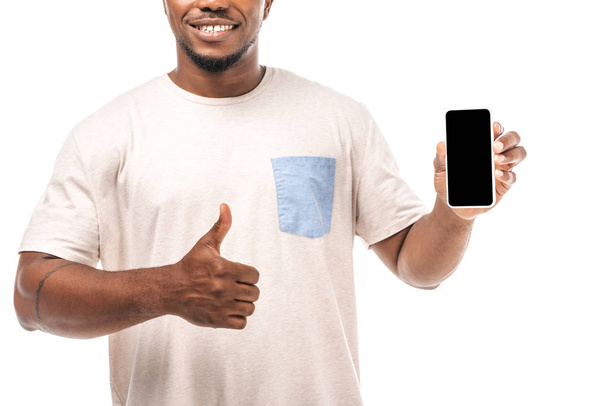 gedeeltelijke weergave van glimlachende Afrikaanse Amerikaanse man met smartphone met leeg scherm en weergegeven duim omhoog geïsoleerd op wit - Foto, afbeelding