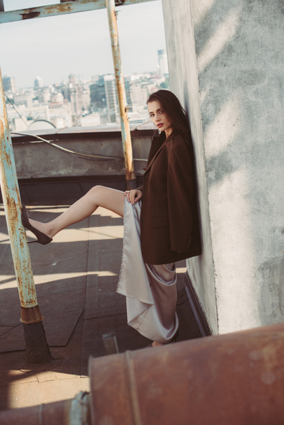 beautiful young woman posing in silk dress and brown jacket on roof - Foto, Bild
