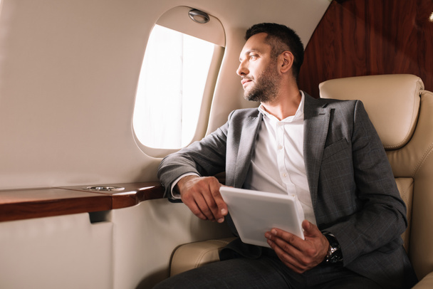 bearded businessman in suit holding digital tablet while looking at airplane window in private jet  - 写真・画像