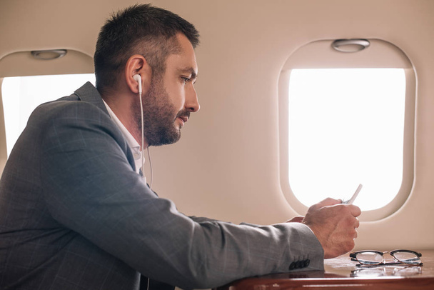 side view of businessman in earphones using smartphone in private jet  - Foto, imagen
