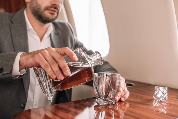 καλλιεργημένη άποψη του γενειοφόρου επιχειρηματία ρίχνει ουίσκι σε ποτήρι στο αεροπλάνο  - Φωτογραφία, εικόνα