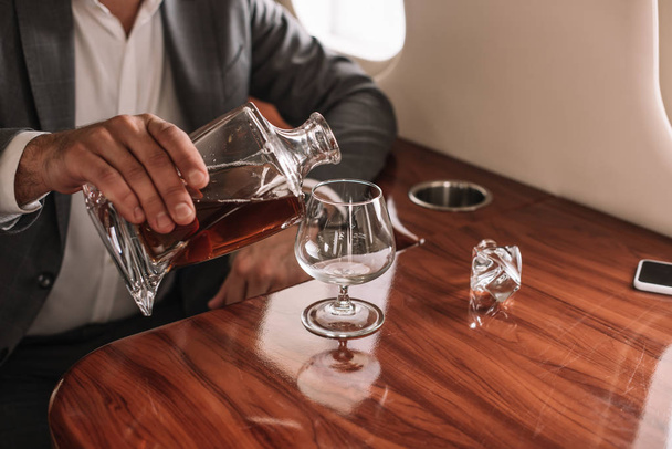 cropped view of businessman pouring cognac into glass in private jet  - Valokuva, kuva