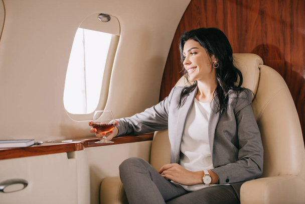 vrolijk zakenvrouw in formele dragen holding glas met cognac in prive jet  - Foto, afbeelding