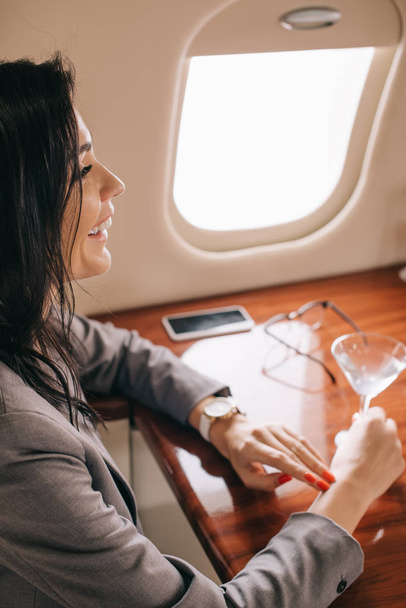 selective focus of cheerful businesswoman holding martini glass in private jet  - Photo, Image