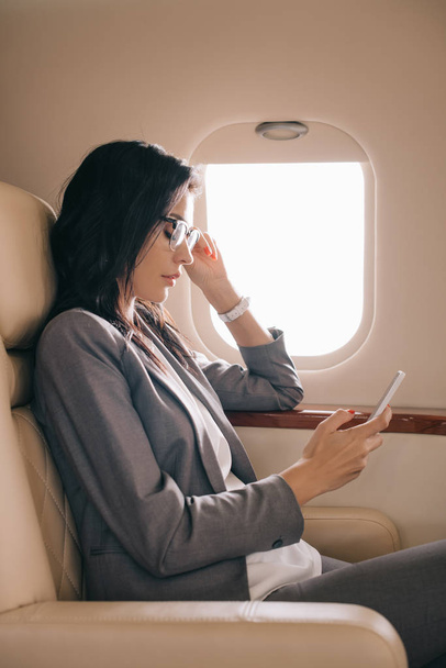 side view of businesswoman in glasses using smartphone in private plane - Foto, immagini
