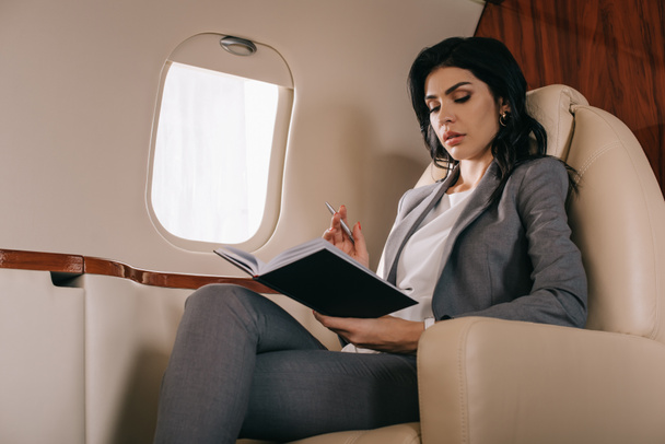 attractive businesswoman holding pen while looking at notebook in private jet  - Foto, afbeelding