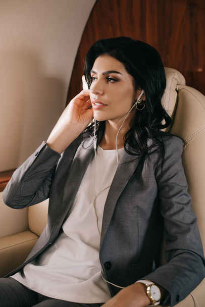 attractive businesswoman touching earphones while listening music in private jet  - Foto, Imagem