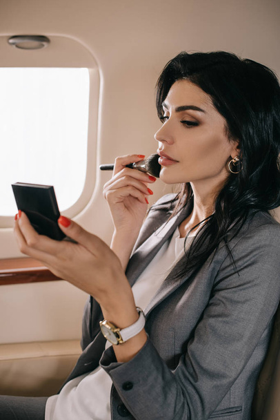 attractive businesswoman holding cosmetic brush near face in private jet  - Fotoğraf, Görsel