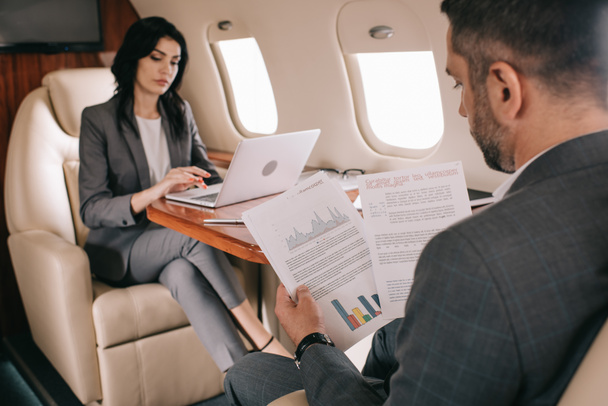 selective focus of bearded businessman holding charts and graphs near attractive businesswoman using laptop in plane  - Фото, изображение