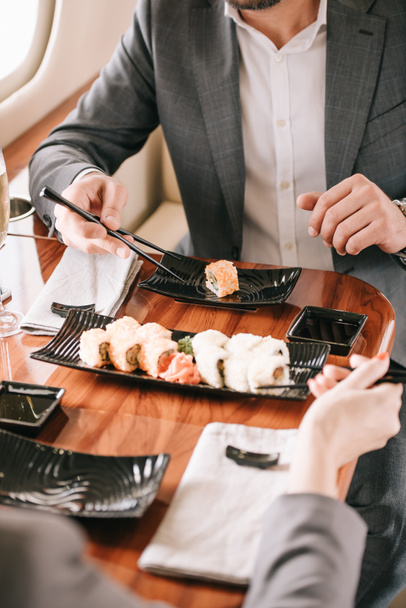 cropped view of businessman and businesswoman holding chopsticks near sushi in private jet  - Foto, Imagen