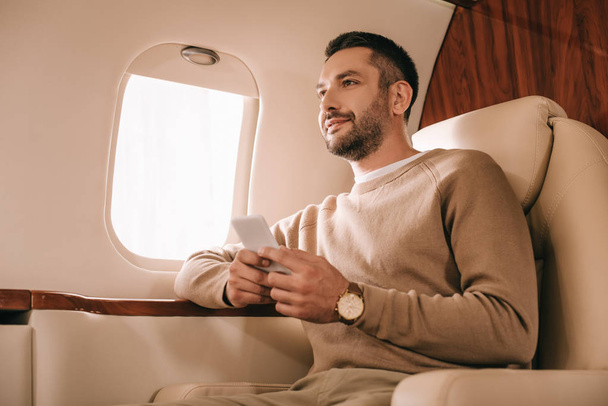 handsome bearded man holding smartphone while sitting in private jet  - Foto, Imagem