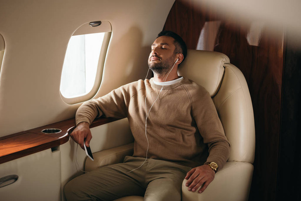 handsome bearded man holding smartphone while listening music in private jet  - Photo, Image