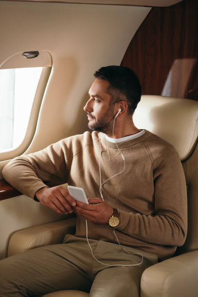 uomo barbuto che tiene smartphone durante l'ascolto di musica in jet privato
  - Foto, immagini