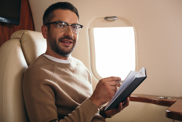 handsome bearded man in glasses looking at camera while holding notebook in private jet  - Foto, Imagem