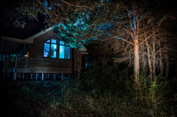 Old house with a Ghost in the forest at night or Abandoned Haunted Horror House in fog. Old mystic building in dead tree forest. Trees at night with moon. Surreal lights. Horror Halloween concept - Photo, Image
