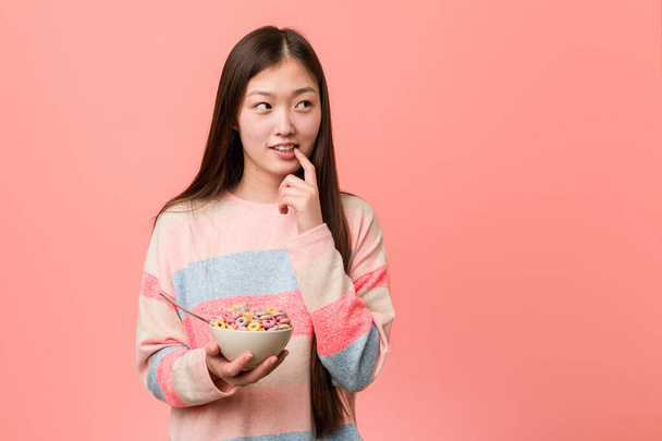 Joven mujer asiática con un tazón de cereal relajado pensando en algo mirando un espacio de copia
. - Foto, imagen