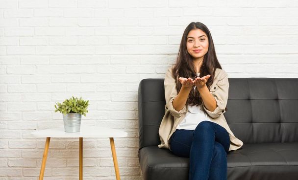Jeune femme arabe assise sur le canapé tenant quelque chose avec des paumes, offrant à la caméra
. - Photo, image