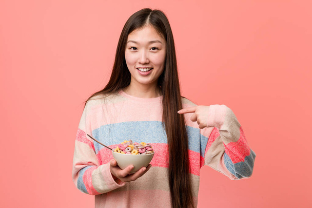 Jovem mulher asiática com uma tigela de cereais surpreso apontando para si mesmo, sorrindo amplamente
. - Foto, Imagem