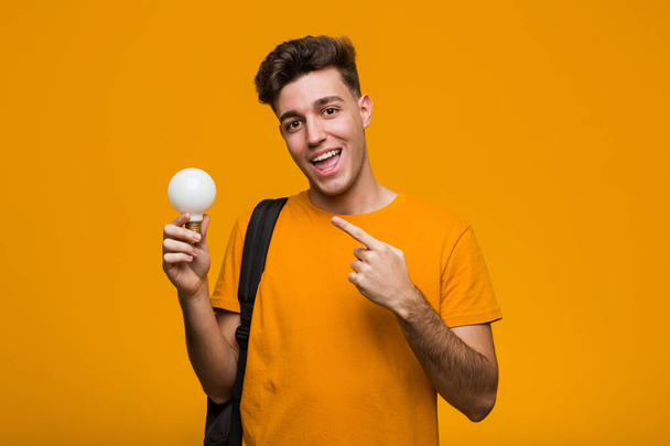Молодий студент тримає лампочку, вказуючи пальцем на вас, ніби запрошений наближається
. - Фото, зображення