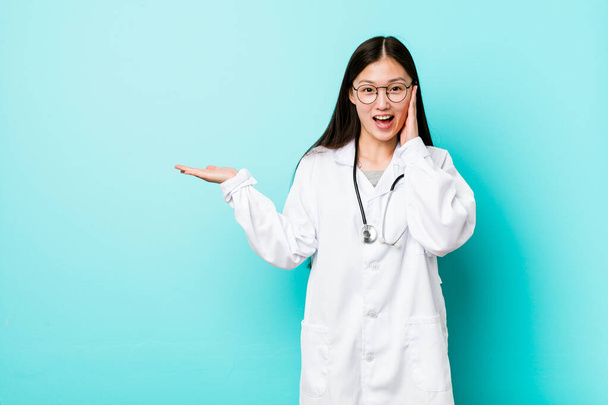 Young chinese doctor woman holds copy space on a palm, keep hand over cheek. Amazed and delighted. - Φωτογραφία, εικόνα