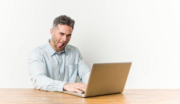 Jonge knappe man werkt met zijn laptop grappig en vriendelijk steken uit tong. - Foto, afbeelding