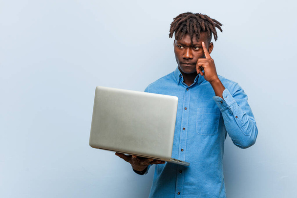 Молодой раста черный человек держит ноутбук указывая пальцем на висок, думая, сосредоточен на задаче
. - Фото, изображение