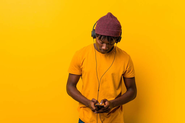 Nuori afrikkalainen mies seisoo keltaista taustaa vasten hattu päässä kuunnellen musiikkia puhelimen kanssa
 - Valokuva, kuva
