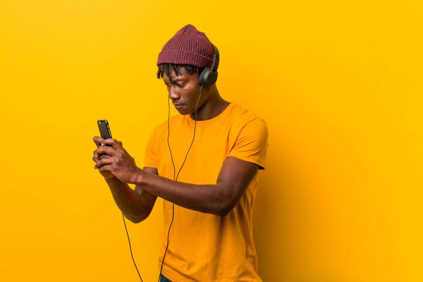 Joven africano de pie sobre un fondo amarillo con un sombrero escuchando música con un teléfono
 - Foto, imagen
