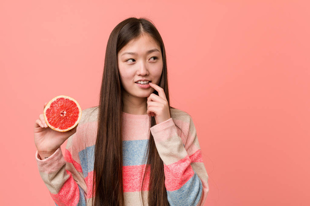 Молода азіатка з грейпфрутом розслабилася думаючи про щось, дивлячись на простір для копіювання
. - Фото, зображення