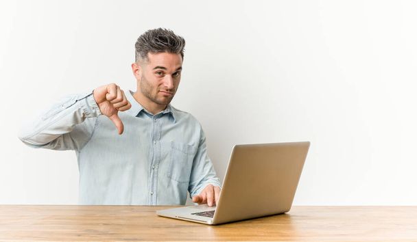Ein junger gutaussehender Mann, der mit seinem Laptop arbeitet und eine ablehnende Geste zeigt, drückt den Daumen nach unten. Unstimmigkeiten. - Foto, Bild