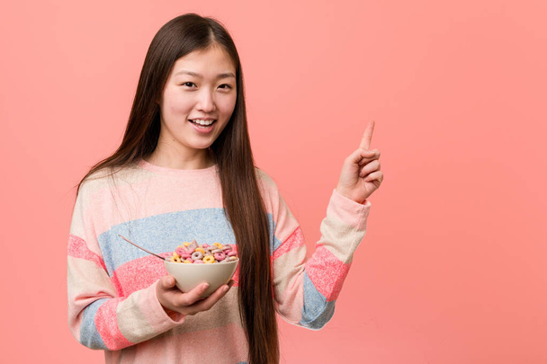 Joven mujer asiática con un tazón de cereal sonriendo alegremente señalando con el dedo índice de distancia
. - Foto, imagen
