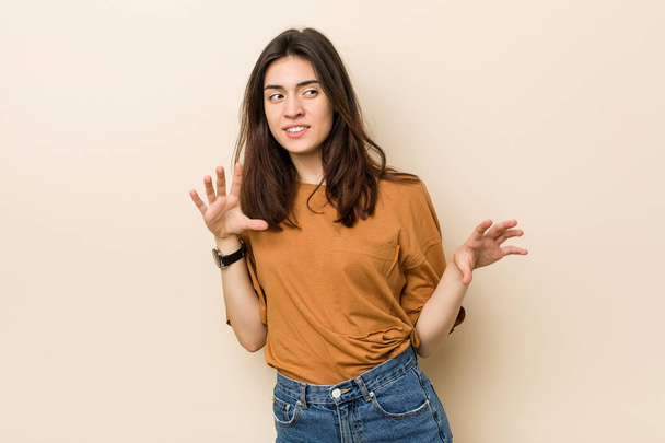 Young brunette woman against a beige background rejecting someone showing a gesture of disgust. - Photo, Image