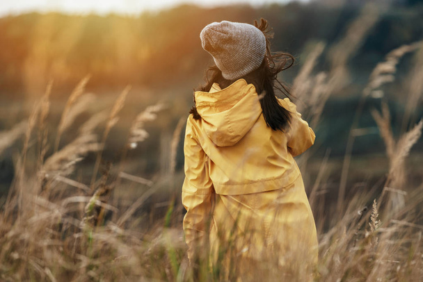Young female with yellow raincoat in tall grass during sunset light - Windy outdoors weather - Фото, изображение