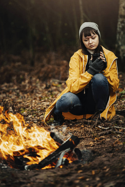 Viajante fêmea em capa de chuva amarela sentada ao lado do fogo do acampamento na floresta com bebida quente na mão / clima de outono escuro na floresta
. - Foto, Imagem