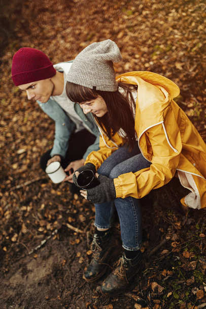 Young happy couple travellers with happy emotions drinking cup of hot drink next to camp fire in forest/ Dark autumn mood in the woods. - Photo, image