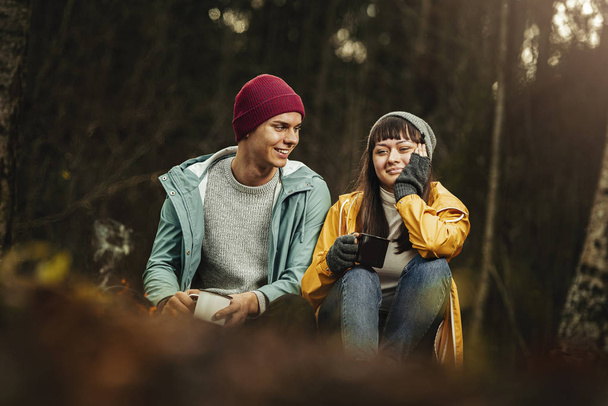 Young happy couple travellers with happy emotions drinking cup of hot drink next to camp fire in forest/ Dark autumn mood in the woods. - Photo, Image