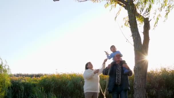 happy kid with grandparents throws rope on tree to make swing on nature in sunny weather - Video