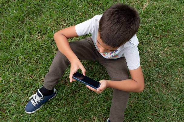 Little boy sitting on the grass using mobile phone in horizontal position. Directly above shot. - Photo, Image