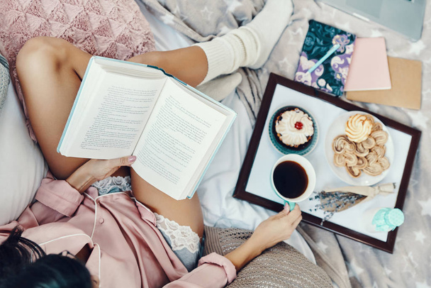  Top view of beautiful young woman in pajamas reading book and enjoying morning coffee while resting in bed at home                 - Foto, imagen