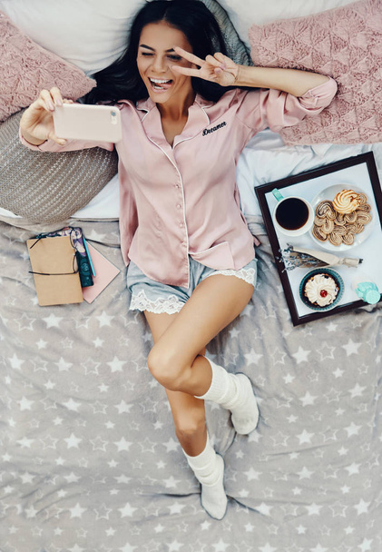 Top view of beautiful young woman in pajamas taking selfie and smiling while resting in bed at home    - Fotografie, Obrázek