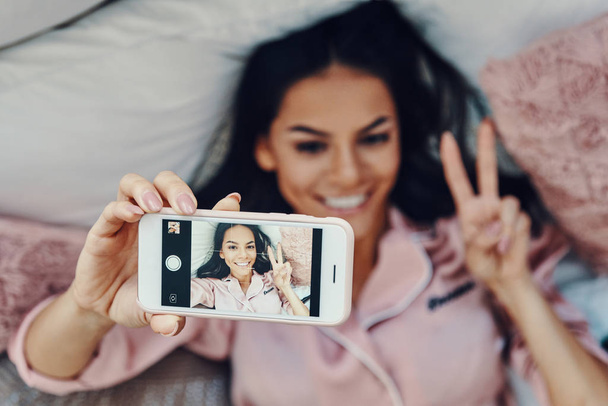 Top view of beautiful young woman in pajamas taking selfie and smiling while resting in bed at home                - Foto, Bild