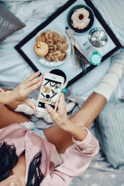 Close up top view of young woman in pajamas photographing flat lay of breakfast while resting in bed at home - Zdjęcie, obraz