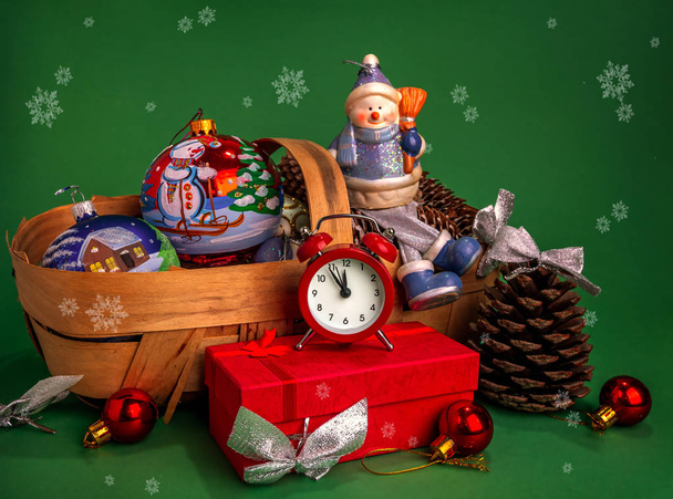Gelukkig Nieuwjaar en Vrolijk Kerstfeest. Sneeuwman, rode geschenkdoos, kerstspeelgoed in een mandje en een grote bult op een groene achtergrond met sneeuwvlokken. - Foto, afbeelding