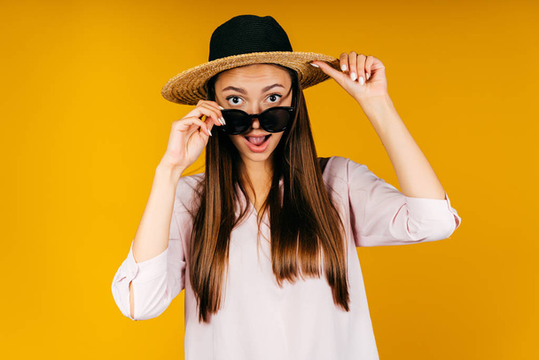 yellow background. girl opens her mouth with her tongue and looks over the glasses in a hat - Foto, Imagen