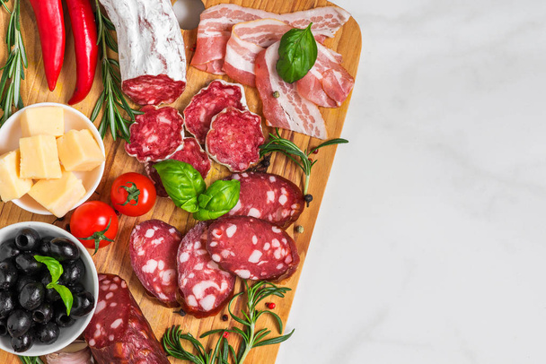 Cutting board with french salami, smoked sausage, bacon, cheese, olives and herbs on white marble background - 写真・画像