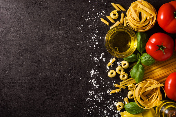 Italiaanse pasta en ingrediënten. Ravioli, penne pasta, spaghetti, tortellini, tomaten en basilicum op zwarte achtergrond. Bovenaanzicht kopieerruimte - Foto, afbeelding