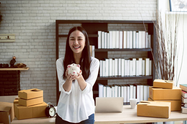 Portrét usměvavé asijské mladé ženy s prasátko banky a lepenkové krabice stojící v kanceláři domu, spuštění on-line marketing koncept - Fotografie, Obrázek