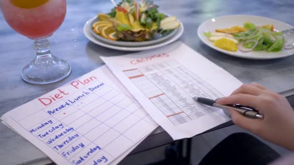 Ernährung, Ernährungswissenschaftlerin Mädchen sucht in Liste der Früchte und ihre Kalorien in der Nähe Punsch und gesunde vegane Nahrung in Teller im Café - Filmmaterial, Video