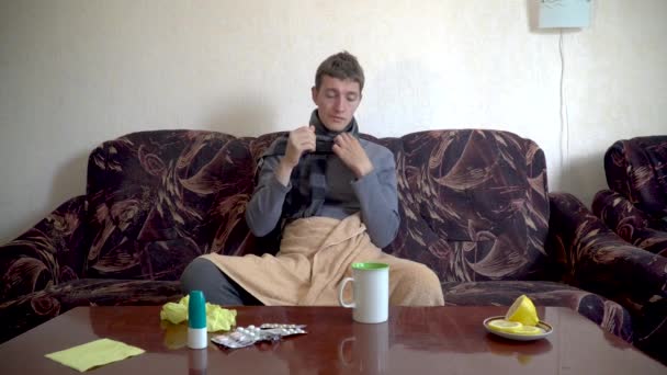 zieke blanke man zit op de bank onder de deken, meet zijn temperatuur en drinkt koude pillen - Video