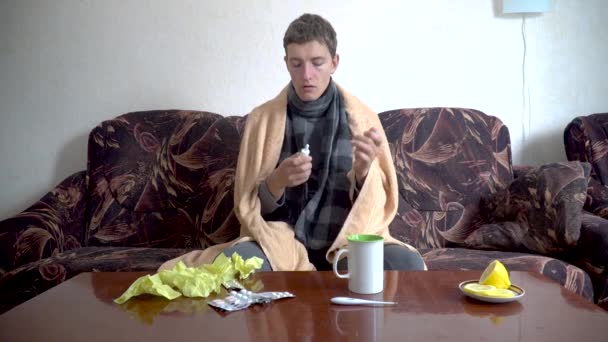 caucasian sick man sits on sofa and uses nasal spray for cold during illness wrapped in blanket - Footage, Video